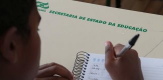 aluno escrevendo com caneta em caderno sobre carteira onde está escrito "secretaria estadual de educação"