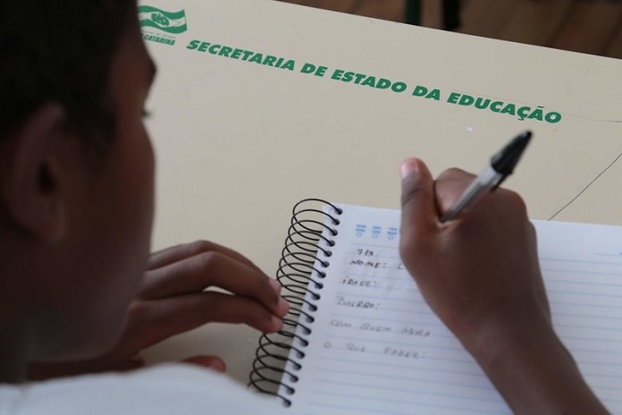 aluno escrevendo com caneta em caderno sobre carteira onde está escrito "secretaria estadual de educação"