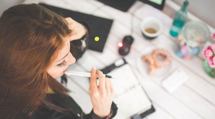 moça vista de cima com caneta na boca em frente à mesa com café e objetos de estudo