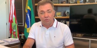 gean fala gesticulando para a câmera em seu gabinete, com bandeira ao fundo de sc, brasil e municipal. ele usa uma camisa polo branca e relógio