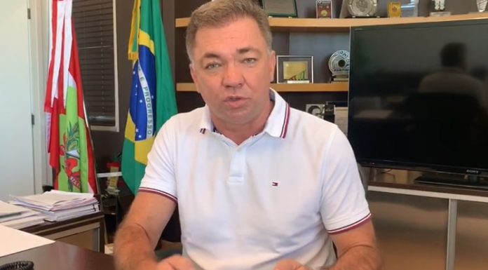 gean fala gesticulando para a câmera em seu gabinete, com bandeira ao fundo de sc, brasil e municipal. ele usa uma camisa polo branca e relógio