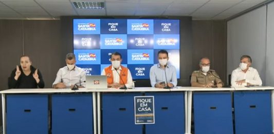 Governo estadual prorroga quarentena até 13/4 e libera comércio automotivo bancada com cinco homens usando máscaras N95 e uma intérprete de libras sem máscara; painel ao fundo com logos do governo de sc