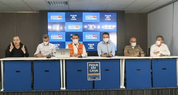 bancada com cinco homens usando máscaras N95 e uma intérprete de libras sem máscara; painel ao fundo com logos do governo de sc