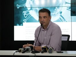 zeferino fala ao microfone com outros microfones de imprensa sobre bancada e telão ao fundo