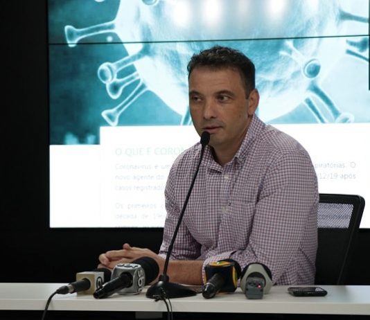zeferino fala ao microfone com outros microfones de imprensa sobre bancada e telão ao fundo