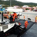 operário fazem obra de asfalto: em primeiro plano uma grande máquina que derrama o asfalto e demais em volta no chão, carros passando ao fundo