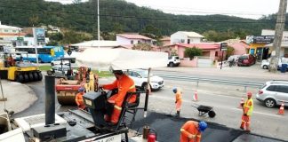 operário fazem obra de asfalto: em primeiro plano uma grande máquina que derrama o asfalto e demais em volta no chão, carros passando ao fundo