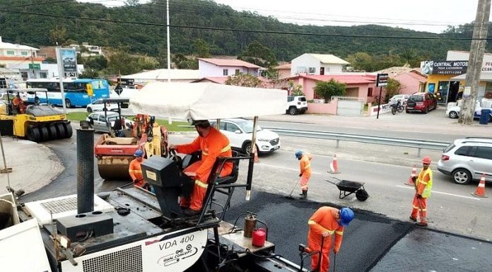 operário fazem obra de asfalto: em primeiro plano uma grande máquina que derrama o asfalto e demais em volta no chão, carros passando ao fundo