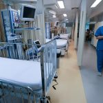 profissional de saúde andando no corredor de uti com leitos expostos pelas cortinas recolhidass