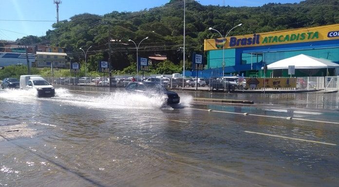 carros passando e espirrando água em rua alagada com loja de supermercado ao fundo