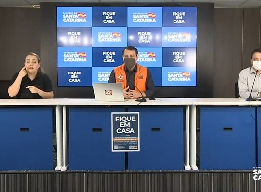 moisés, zeferino e a intérprete de libras em bancada com microfones; eles estão de máscara; a mulher não; painel eletrônico ao fundo com logos do governo