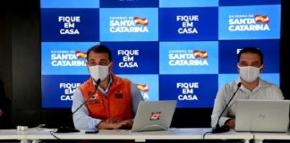 Carlos Moisés e Helton Zeferino em coletiva de imprensa online usando máscaras; painel de logos do governo ao fundo e mensagem "fique em casa"