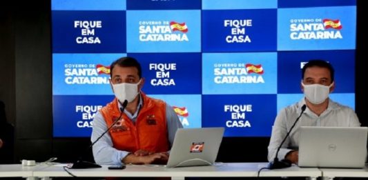 SC tem 37 óbitos por Covid-19 Carlos Moisés e Helton Zeferino em coletiva de imprensa online usando máscaras; painel de logos do governo ao fundo e mensagem "fique em casa"