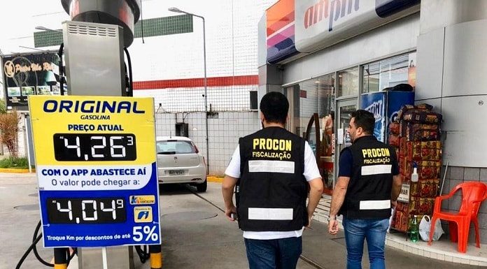 dois fiscais do procon com coletes identificativos entrando em um posto com placa de valor da gasolina de R$4,26