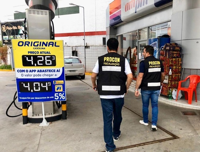 dois fiscais do procon com coletes identificativos entrando em um posto com placa de valor da gasolina de R$4,26