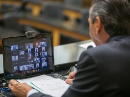 Aprovados projetos sobre recursos do Badesc e prorrogação do ICMS deputado julio garcia com um notebook na frente com transmissão de video dos deputados na tela