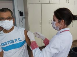 mulher profissional de saúde usando máscara e jaleco aplica vacina no braço de um senhor com camisa do santos fc