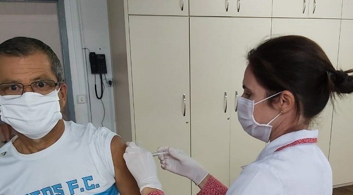 mulher profissional de saúde usando máscara e jaleco aplica vacina no braço de um senhor com camisa do santos fc