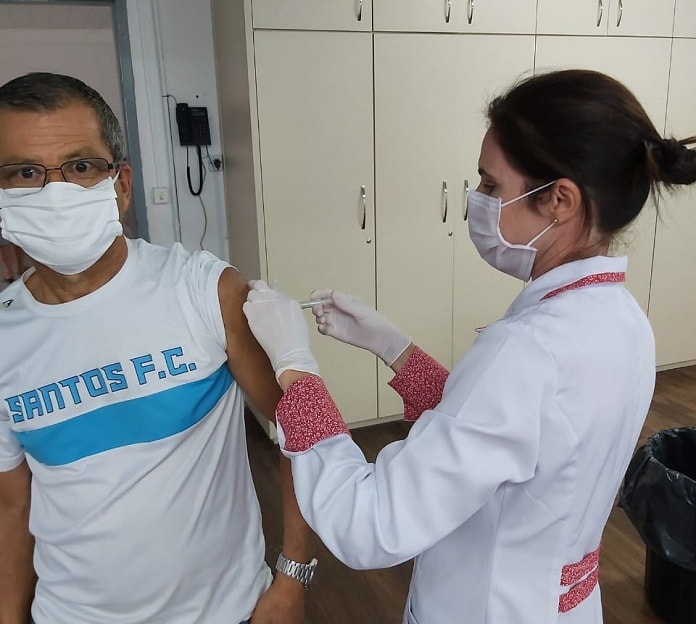 mulher profissional de saúde usando máscara e jaleco aplica vacina no braço de um senhor com camisa do santos fc