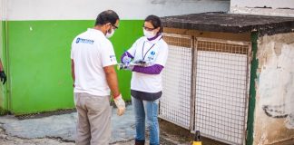 homem e mulher usando equipamentos de proteção e uniformes do programa em pé ao lado de bueiro aberto, com cone, e a mulher aponta algo na prancheta; estão atrás de uma construção