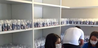 duas mulheres usando máscaras, sentadas,montando kits de proteção individual
