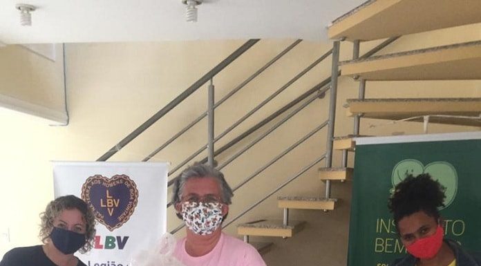 mulheres usando máscaras seguram as cestas básicas ao lado de pilha de pacotes de alimentos