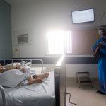profissional de saúde trajado com roupas de proteção e máscara toca em pé violão para senhora deitada em cama de hospital em quarto onde há janela e televisão