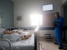 profissional de saúde trajado com roupas de proteção e máscara toca em pé violão para senhora deitada em cama de hospital em quarto onde há janela e televisão