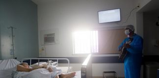 profissional de saúde trajado com roupas de proteção e máscara toca em pé violão para senhora deitada em cama de hospital em quarto onde há janela e televisão