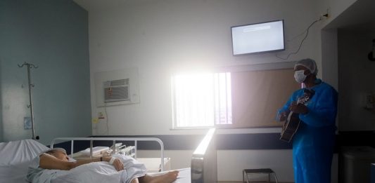 BR tem 182 mil casos ativos de infecção por coronavírus, 2,6 mil em SC profissional de saúde trajado com roupas de proteção e máscara toca em pé violão para senhora deitada em cama de hospital em quarto onde há janela e televisão