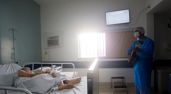 profissional de saúde trajado com roupas de proteção e máscara toca em pé violão para senhora deitada em cama de hospital em quarto onde há janela e televisão