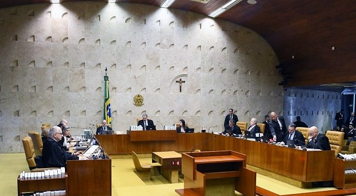 ministros do stf em reunião no plenário da corte; onde há bandeira do brasil e cruz ao fundo na parede