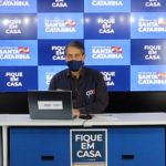 imagem da coletiva onde está, em uma bancada, o secretário ao centro, outro homem ao lado, e intérprete de libras do outro; painel de logos do governo ao fundo