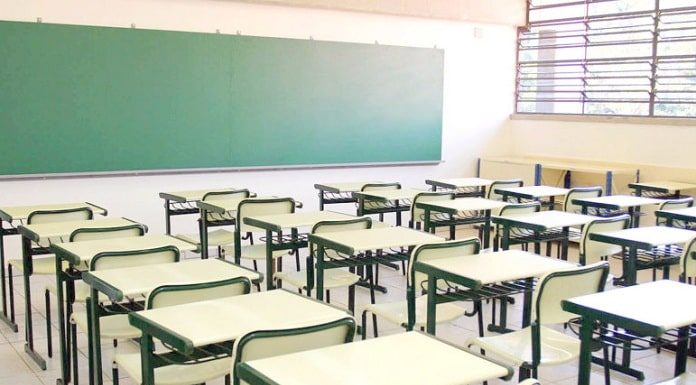 sala de aula vazia com mesas e cadeiras organizadas em fileiras; quadro negro à frente