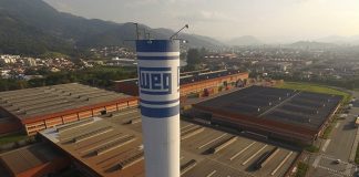 Fábrica da Weg em Jaraguá do Sul vista em foto aérea com torre de caixa d'água com logo da empresa em primeiro plano, galpões em baixo, e cidade ao fundo