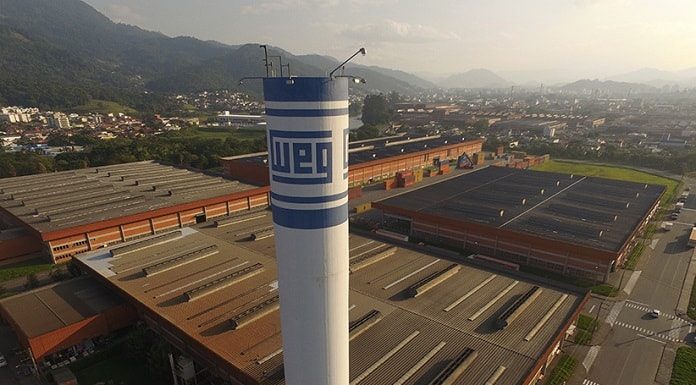 Fábrica da Weg em Jaraguá do Sul vista em foto aérea com torre de caixa d'água com logo da empresa em primeiro plano, galpões em baixo, e cidade ao fundo