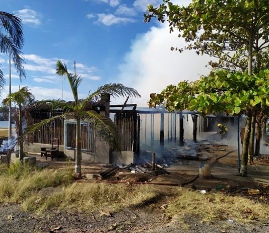 rancho destruído pelo incêndio, com fumaça saindo e algumas árvores em volta; bombeiro ao lado