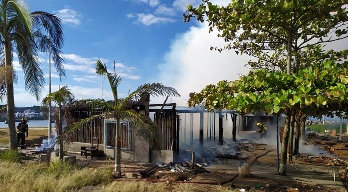 rancho destruído pelo incêndio, com fumaça saindo e algumas árvores em volta; bombeiro ao lado