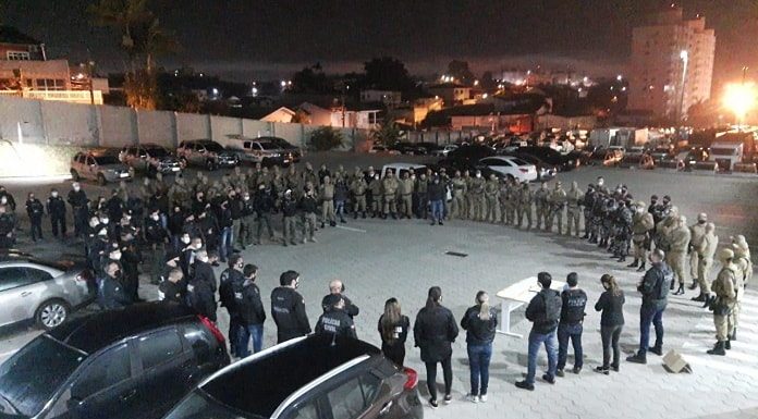 policiais civis e militares em pé em grande roda num estacionamento em foto noturna