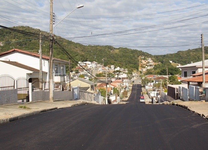 asfalto novo sem pintura em rua que tem leve declive e se estende ao fundo da imagem, com morro no final e casas em volta