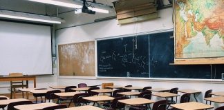 carteiras e mesas de madeira organizadas em sala de aula vazia com quadros negros ao fundo e mapas