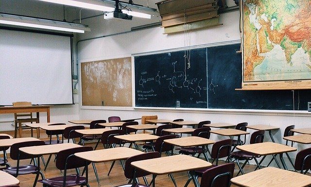 carteiras e mesas de madeira organizadas em sala de aula vazia com quadros negros ao fundo e mapas