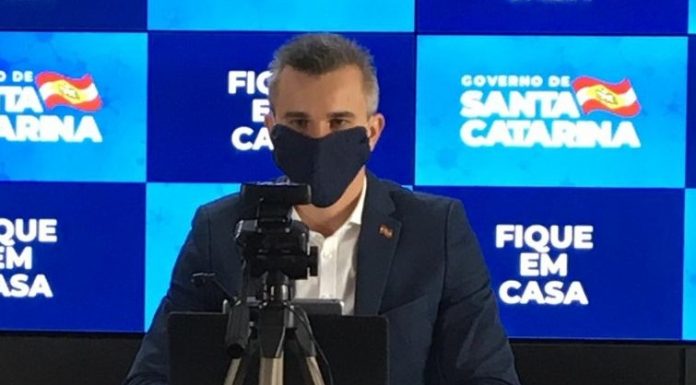 borba usa máscara preta ao dar entrevista online; há uma câmera em tripé na sua frente e painel do governo ao fundo