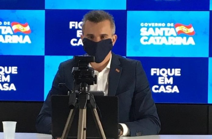 borba usa máscara preta ao dar entrevista online; há uma câmera em tripé na sua frente e painel do governo ao fundo