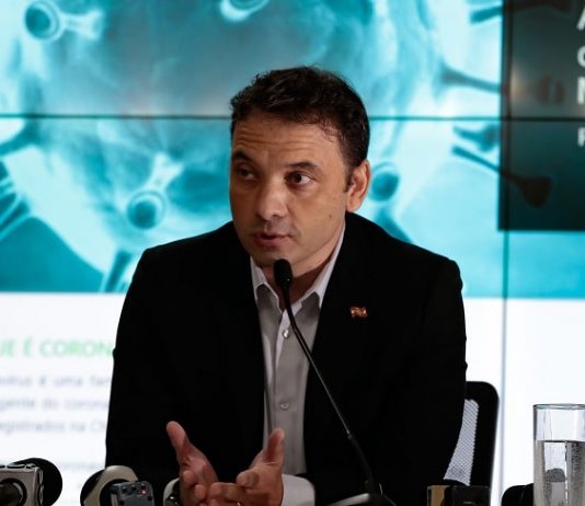 helton zeferino em entrevista; sentado atrás de bancada onde há microfones de imprensa e telão ao fundo com desenhos de coronavírus