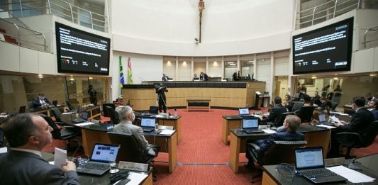 deputados sentados nas bancadas do plenário da alesc, com dois telões ao fundo e mesa diretora, com grande cruz no meio