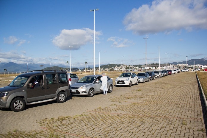carros em fila no estacionamento externo do centro multiuso de são josé; profissional da saúde com trajes de proteção em pé ao lado