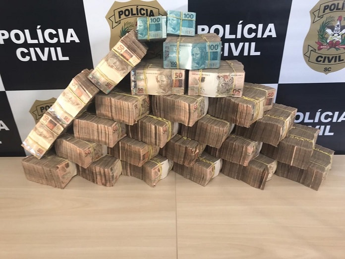 enorme pilha de dinheiro organizado sobre mesa em maços de 50 reais em maioria, na frente de painel da polícia civil