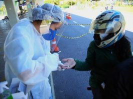 profissional de saúde trajada com equipamentos de segurança espeta dedo de mulher em garupa de moto usando capacete estendendo a mão em um terminal de ônibus