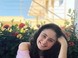 Moça vestindo blusa rosa e calça azul, sentada na calçada com a mão esquerda no cabelo